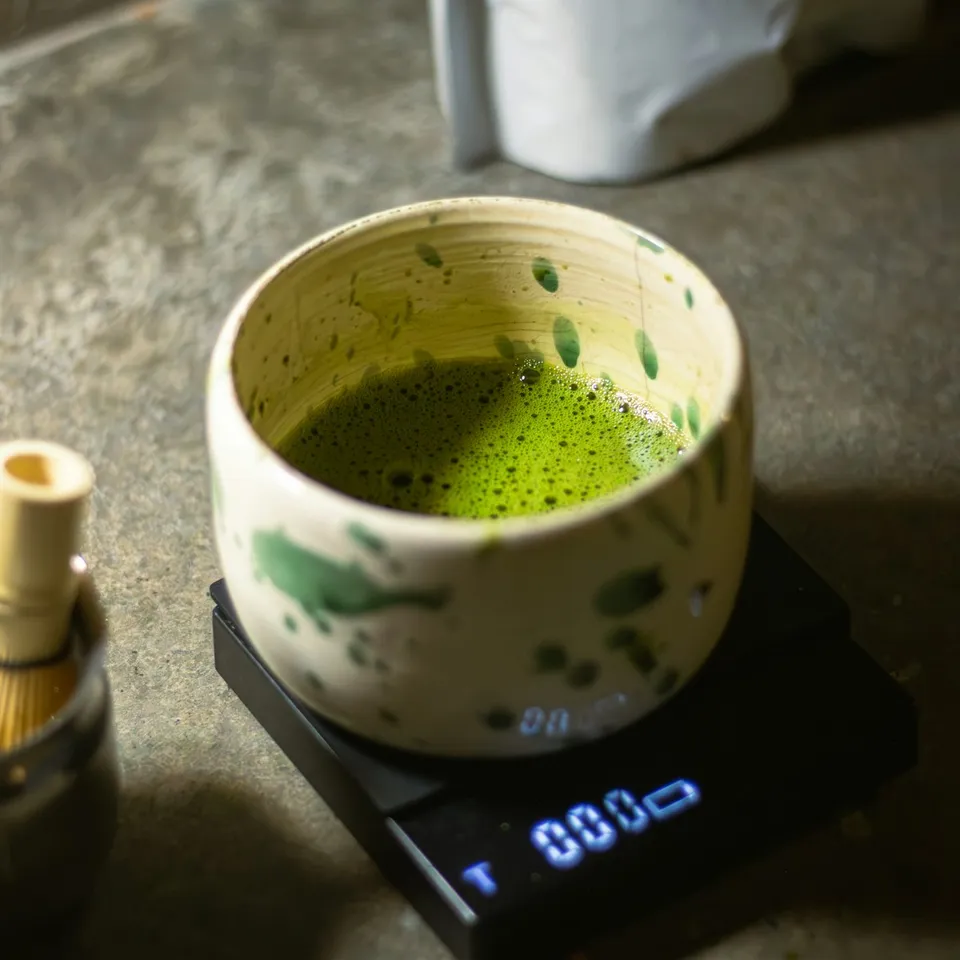 A speckled ceramic bowl of whisked matcha on a kitchen scale, with a bamboo whisk beside it