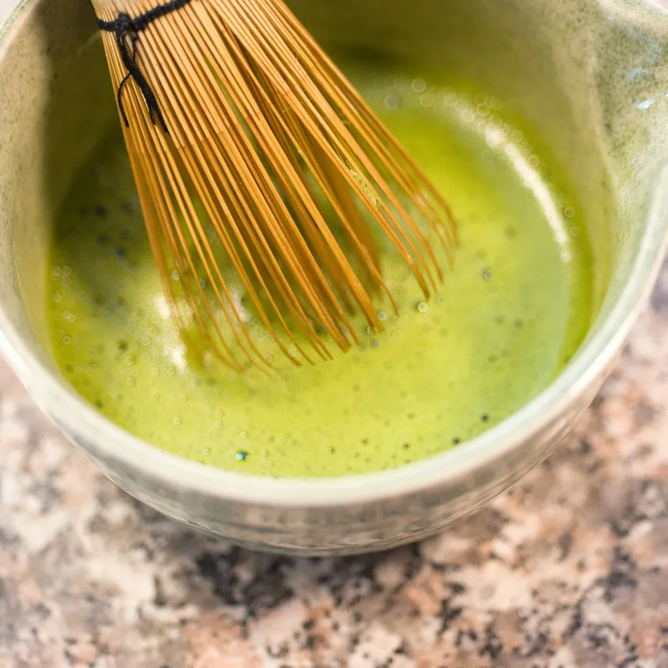 A bamboo whisk resting in a bowl of freshly whisked matcha on a stone counter